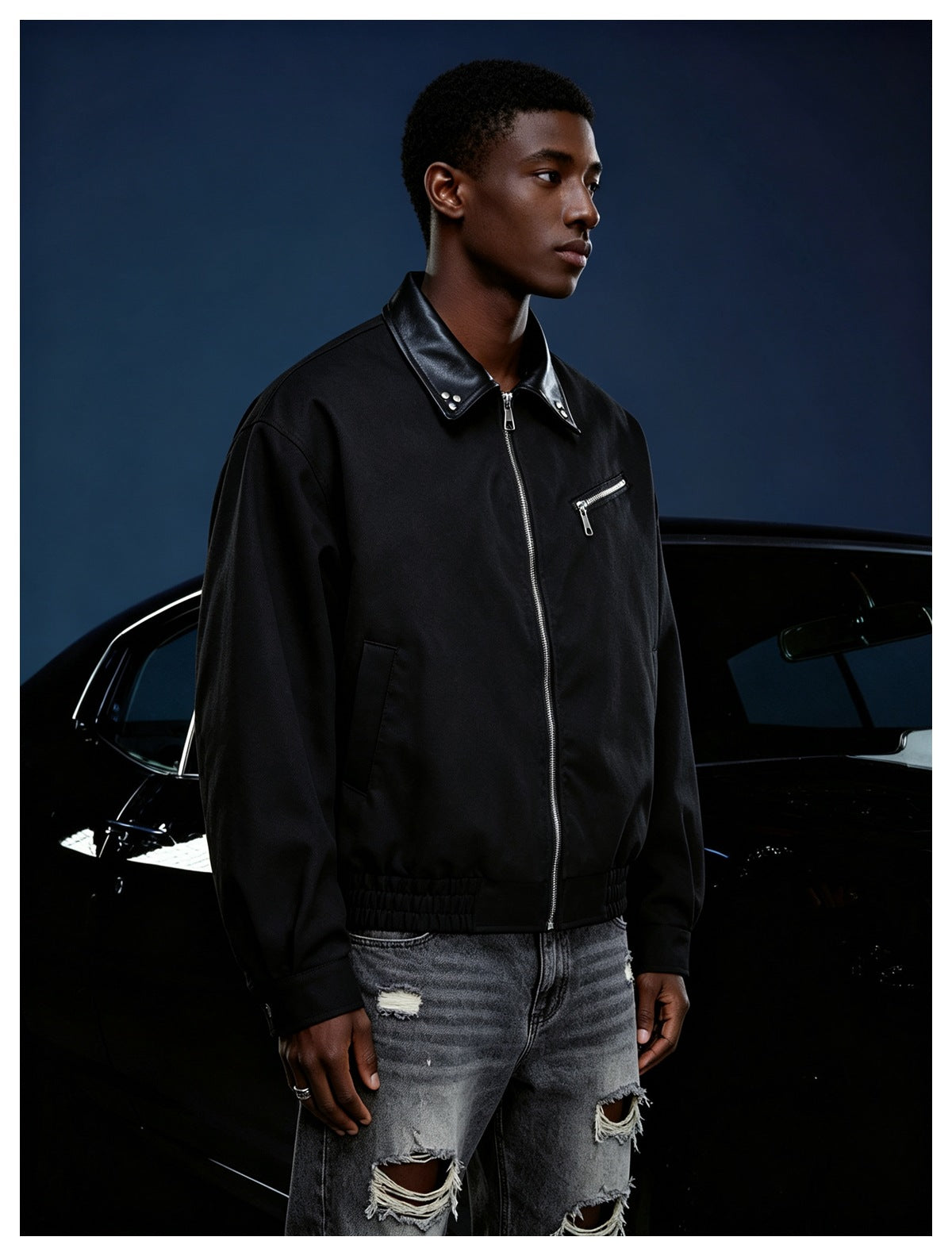 Man wearing black zip-front jacket with studded collar and chest zip pocket, paired with distressed gray jeans