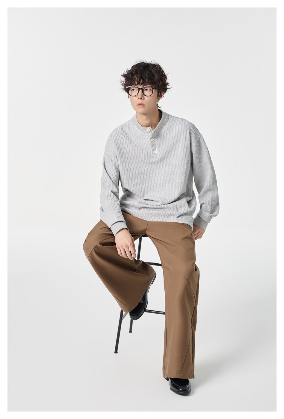 Seated full-length view of a model in a pale gray textured button-neck top, brown trousers, and black shoes.