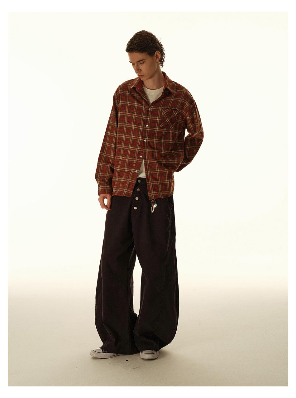 Model looking down in a rust plaid button-up shirt, cream tee, and oversized black trousers.
