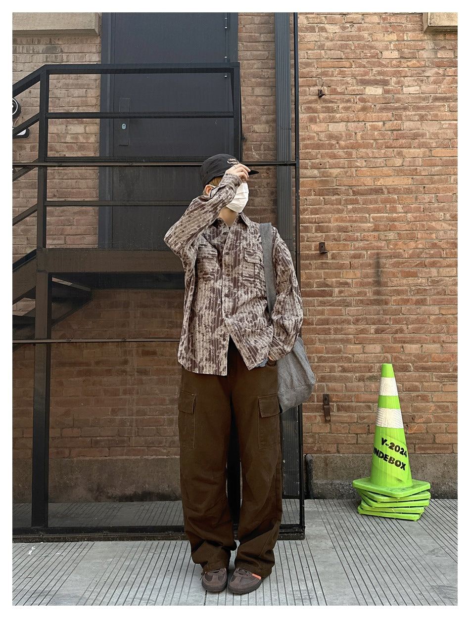 Person in brown striped tie-dye-style shirt with brown cargo pants, standing by a brick wall with arm raised to cap