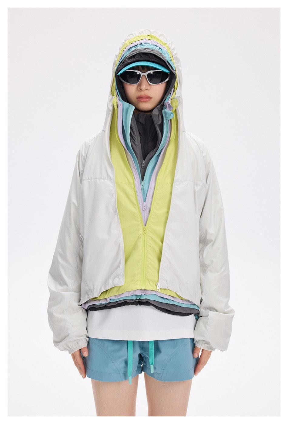 Model wears a white jacket over layered lime, lavender, aqua, and charcoal hooded layers, with blue shorts.
