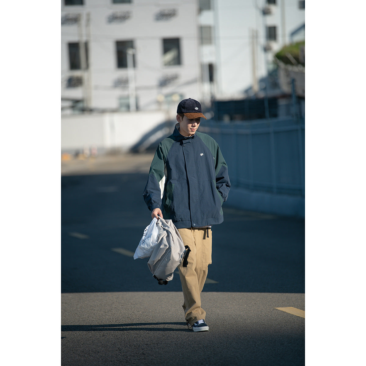Person wearing a zipped navy windbreaker with dark green shoulder panels and cream accents, tan pants and a dark cap.