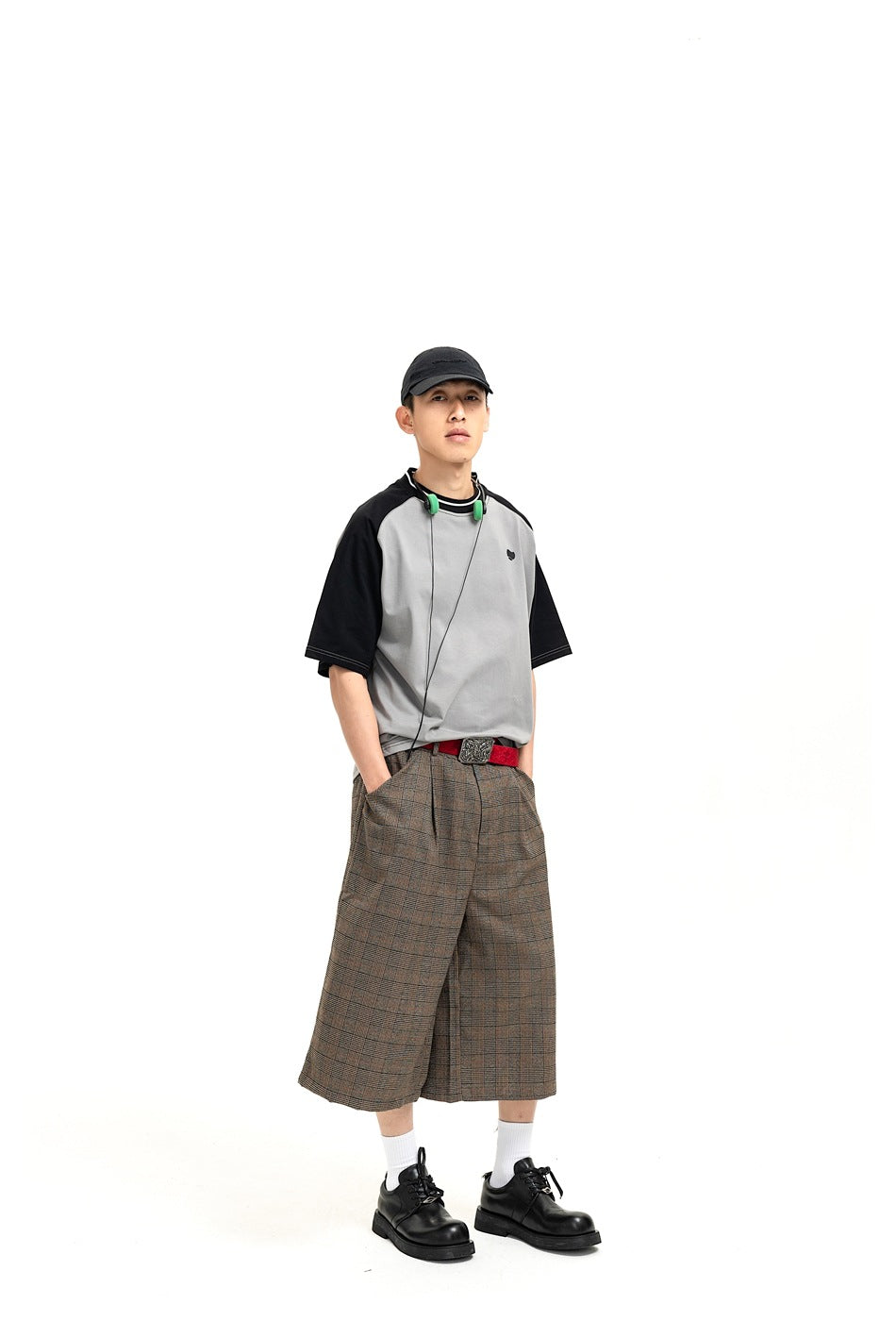 Man wearing gray and black raglan T-shirt with plaid wide-leg cropped shorts, black cap and black shoes, front view