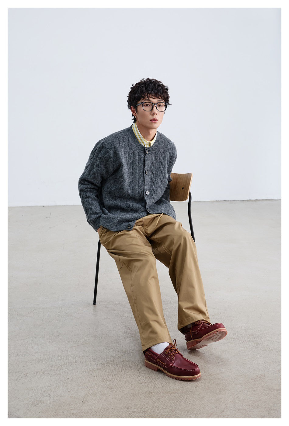 Seated model wearing a charcoal gray cable-knit button cardigan, tan trousers, and burgundy shoes.