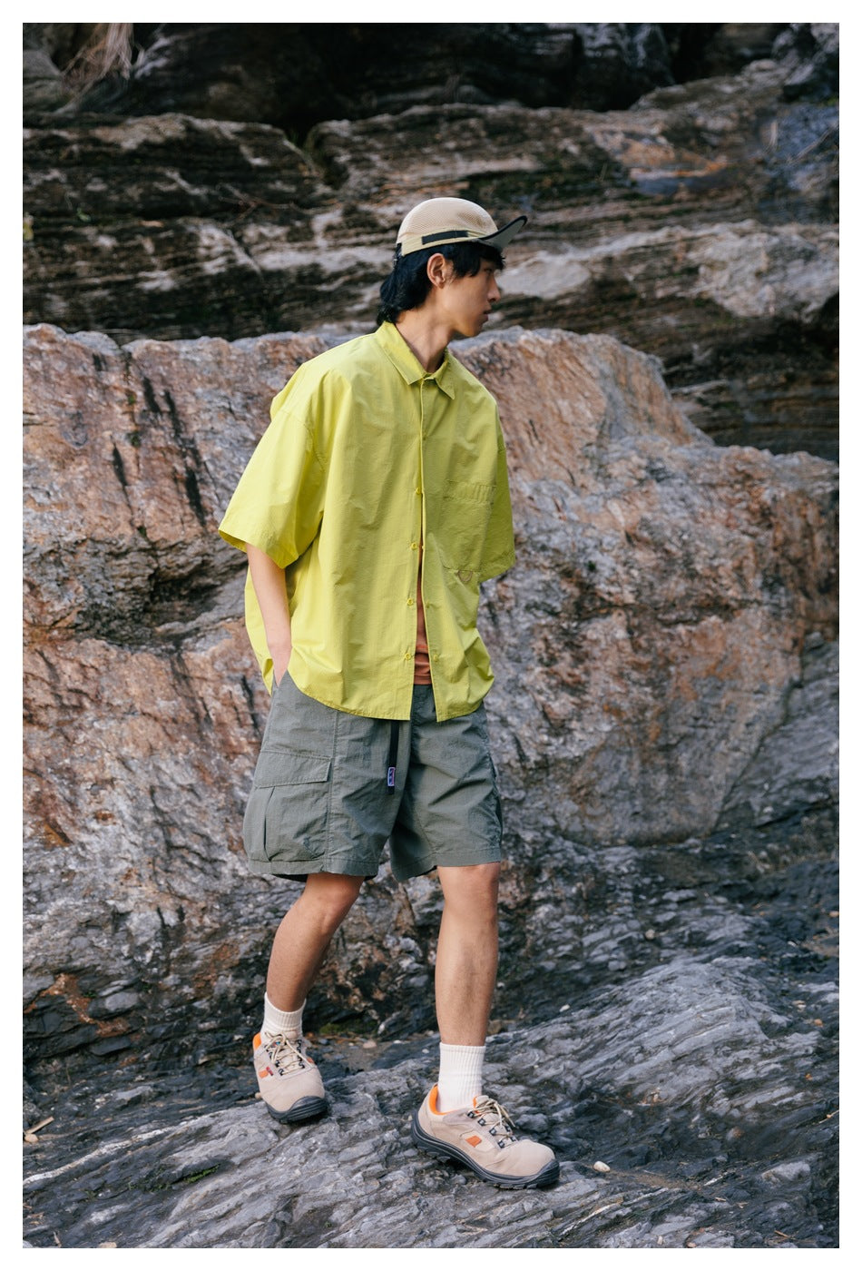A man wears a lime-yellow short-sleeve button-up shirt and gray cargo shorts among rocky cliffs.
