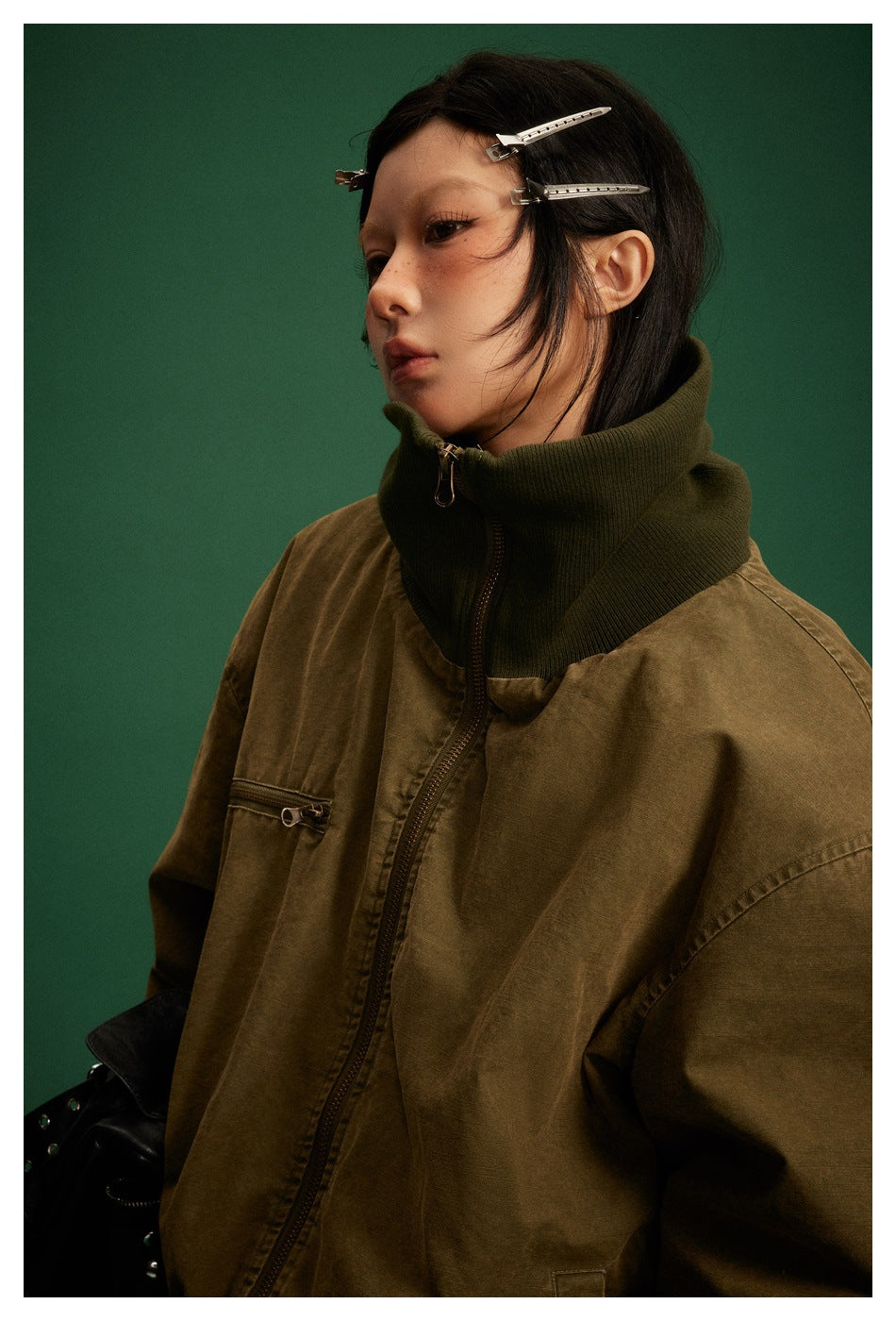 Close view of a model wearing an olive-brown zip jacket with a tall dark ribbed collar and chest zip pocket.