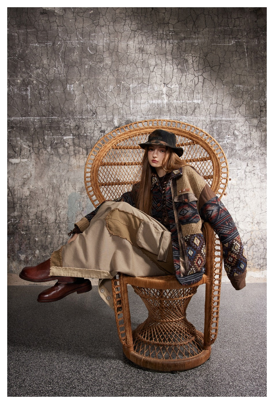 Model seated in a wicker chair wearing a patchwork jacket with geometric patterns, beige trousers, and a dark hat.