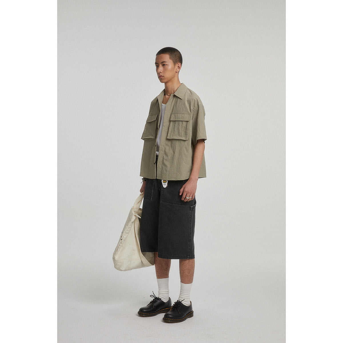 Model wearing an olive short-sleeve button-up shirt with large flap pockets and black knee-length shorts.