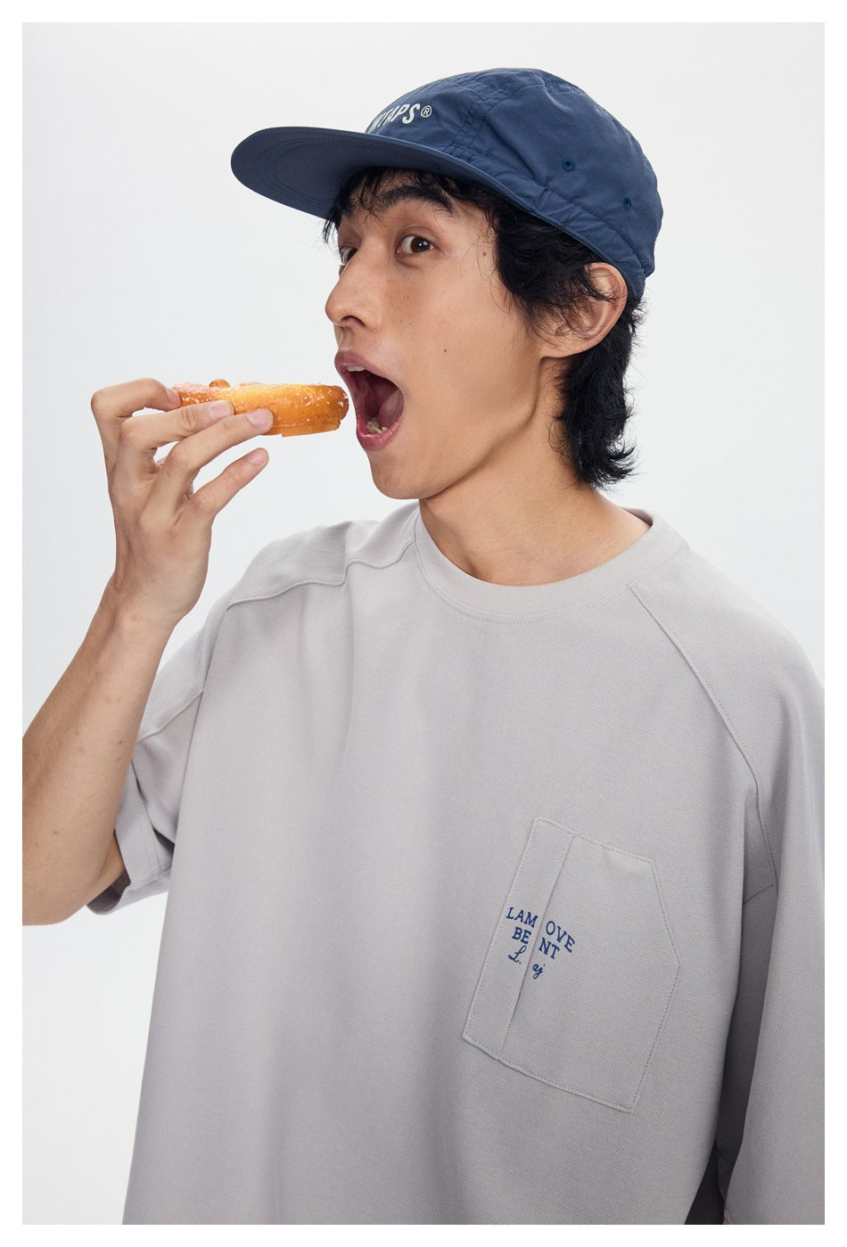 Close view of a model in a light gray pocket T-shirt and navy cap, holding a snack near his mouth.