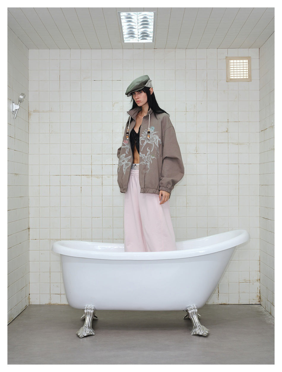 Model wearing a taupe graphic jacket and pale pink pants, posed standing in a white bathtub.