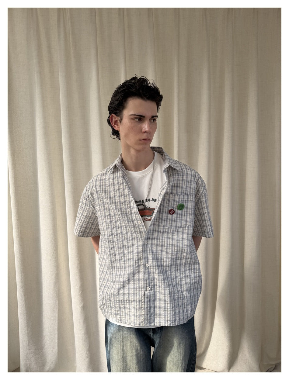 Front view of a model wearing a white and blue plaid short-sleeve shirt with a chest pocket and two pins.