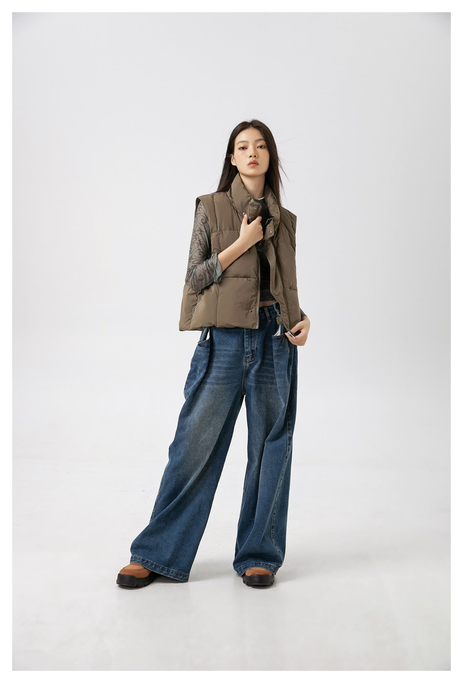 Full-length model wearing a taupe padded vest, patterned sheer long-sleeve top, wide-leg blue jeans and dark shoes.
