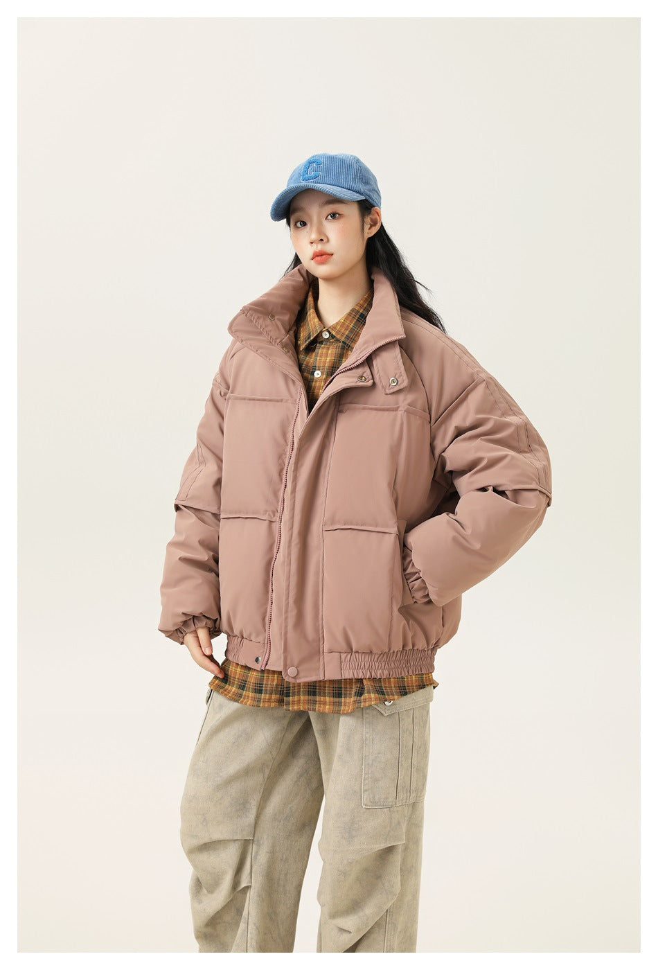 Model wearing a dusty pink puffer jacket over a plaid shirt with a blue cap and light cargo pants, front view