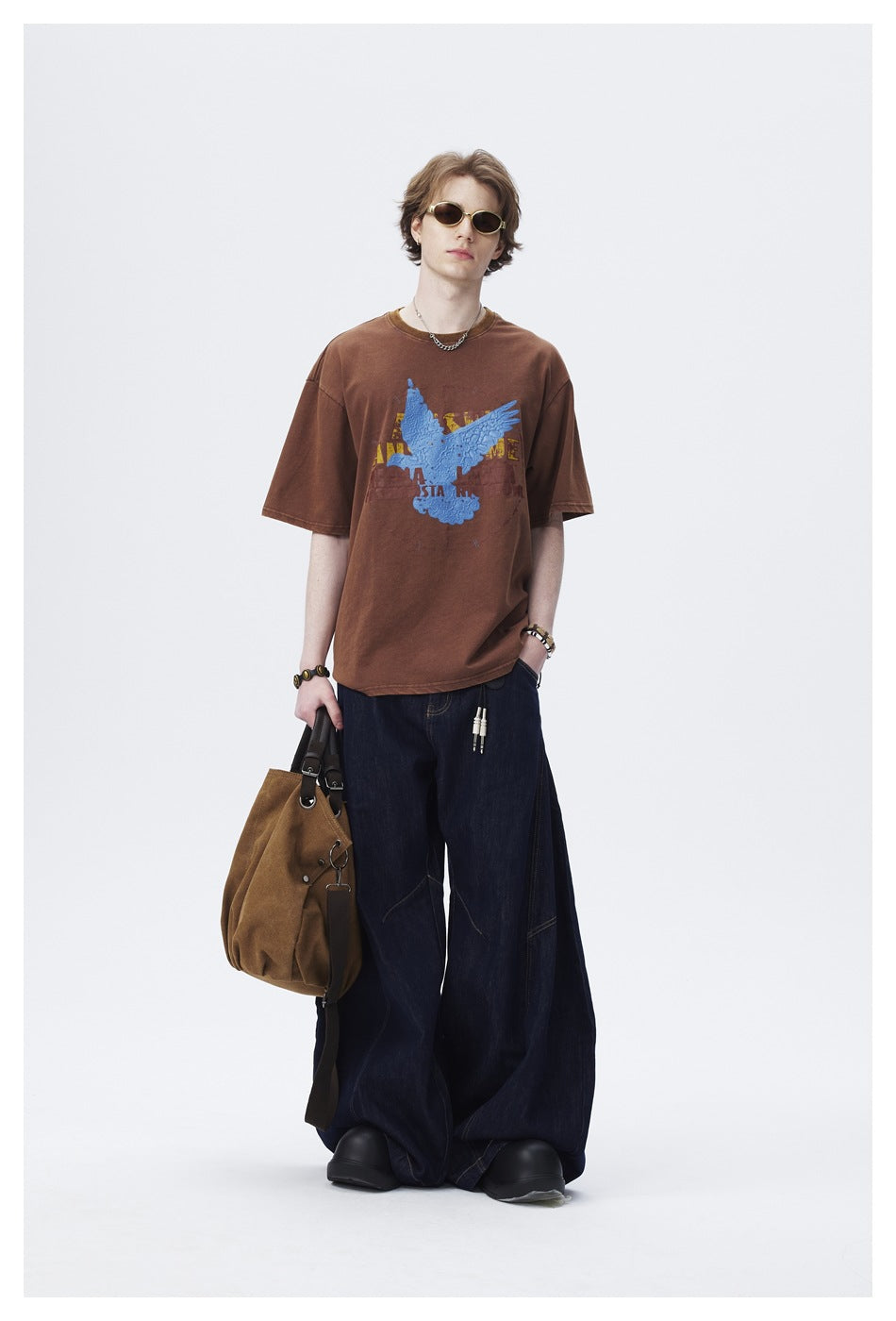 Model in brown bird-graphic T-shirt with wide dark denim jeans, sunglasses and a brown canvas tote bag