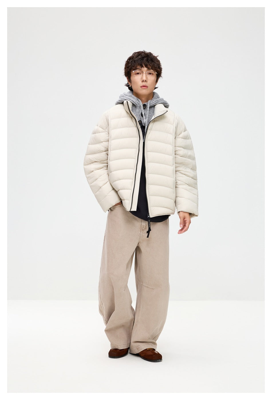 Man wearing a cream quilted puffer jacket over a gray hooded knit with wide beige trousers, front view
