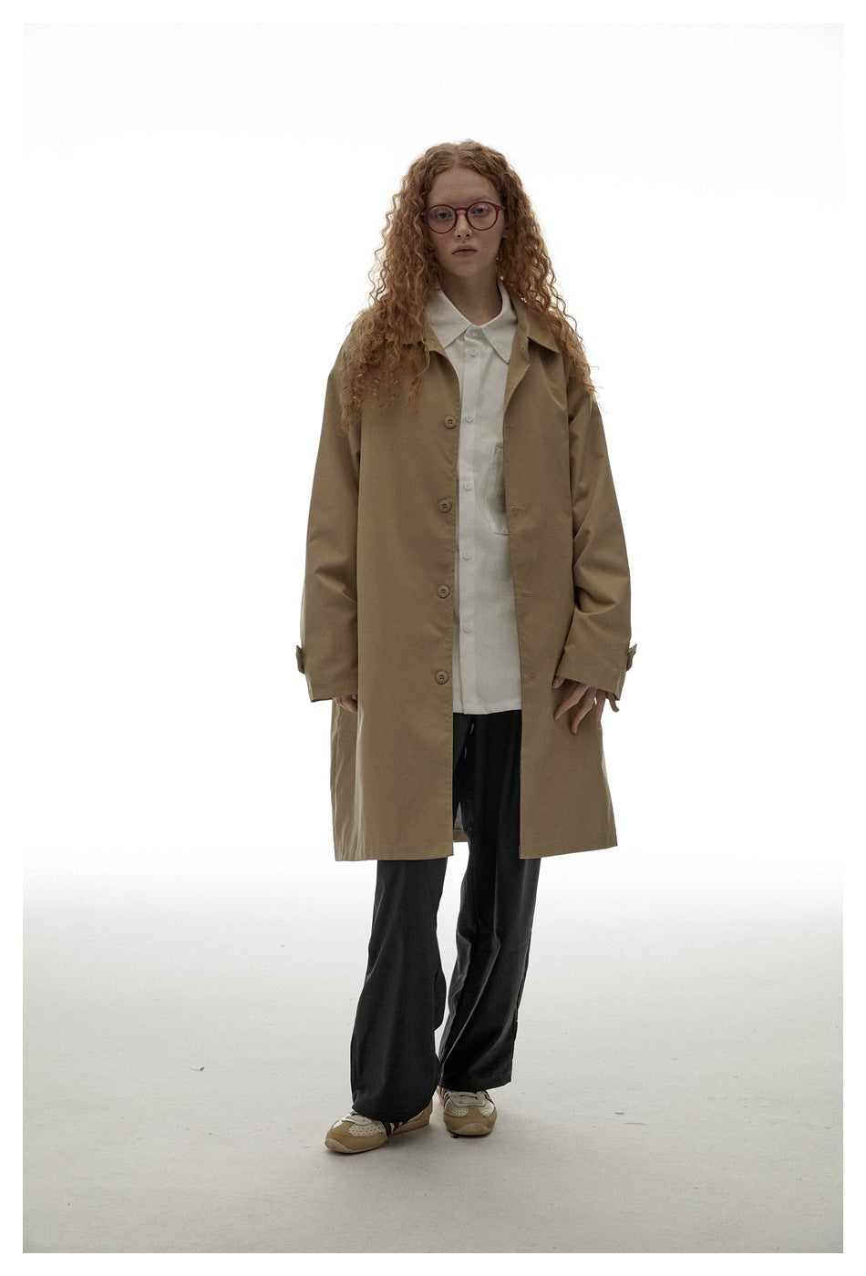 Full-length model wearing a tan button-front coat with a white shirt, black trousers and red glasses.