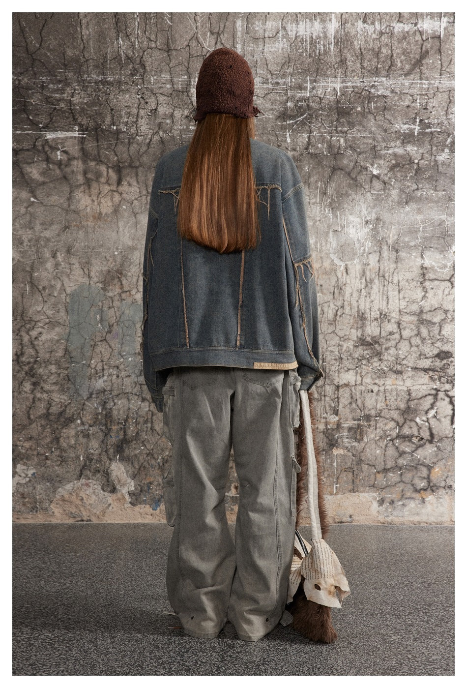 Back view of a model wearing an oversized distressed blue denim jacket and loose gray cargo pants.