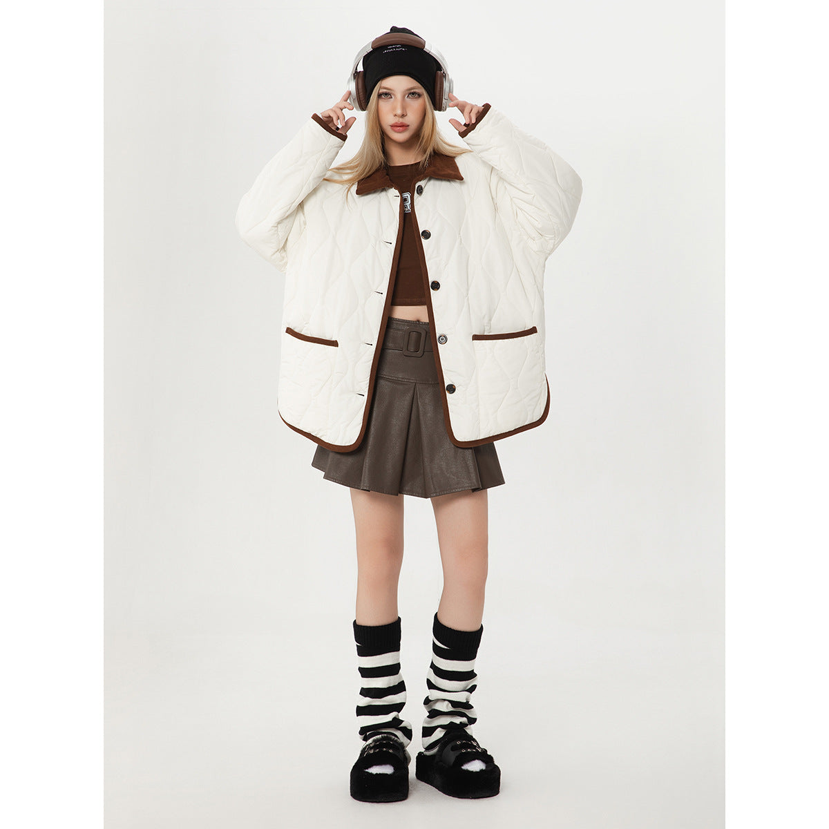 Model wearing a white quilted jacket with brown trim, collar and buttons, styled with a brown skirt and striped leg warmers.
