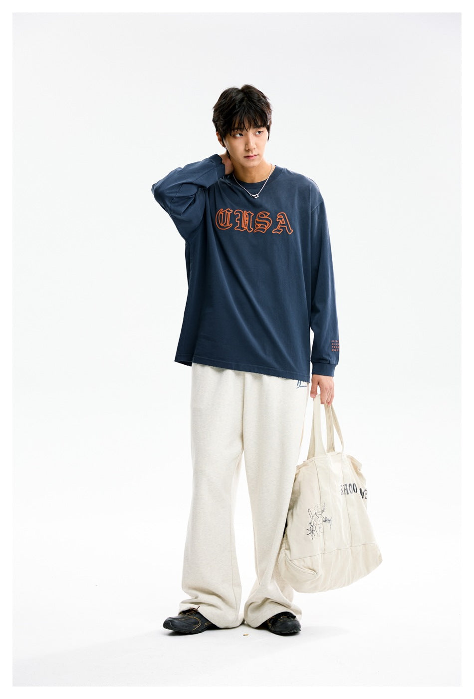 Full-length model wearing a navy USA graphic shirt and cream wide-leg pants, with a canvas tote.