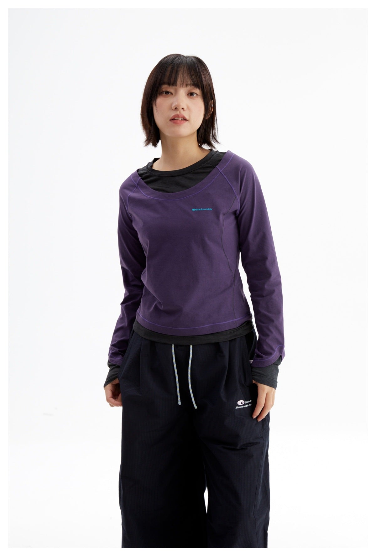 Model wearing a purple long-sleeve top over a black layer and loose black pants, shown from the front.