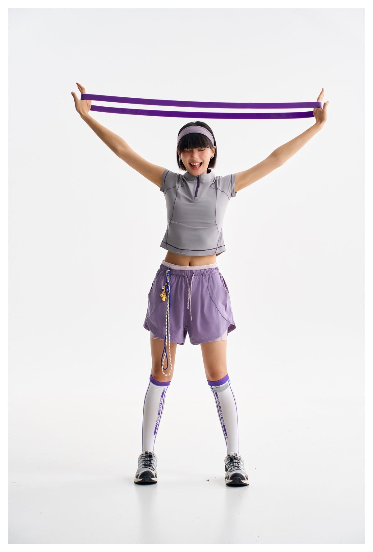 Model in a gray-lavender zip-neck top and purple shorts stretching a purple resistance band overhead.