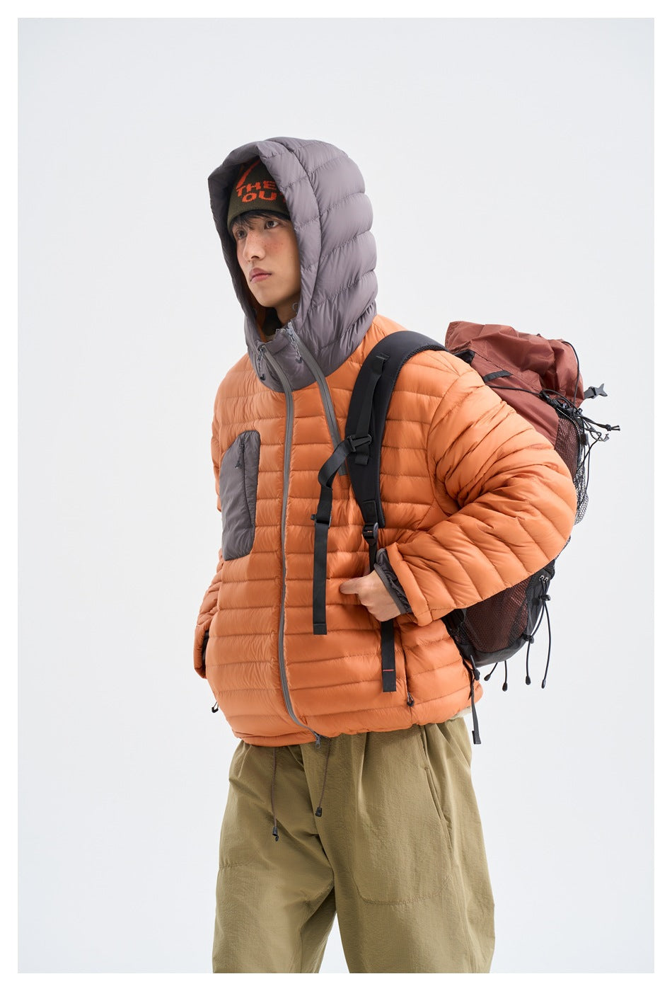 Model wearing an orange puffer jacket with a gray hood and carrying a brown hiking backpack.