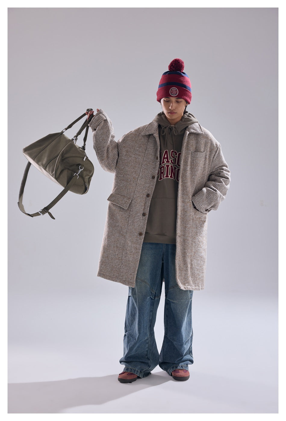 Model wearing a beige patterned long coat over a hoodie and jeans, with a red striped beanie and olive duffel bag.