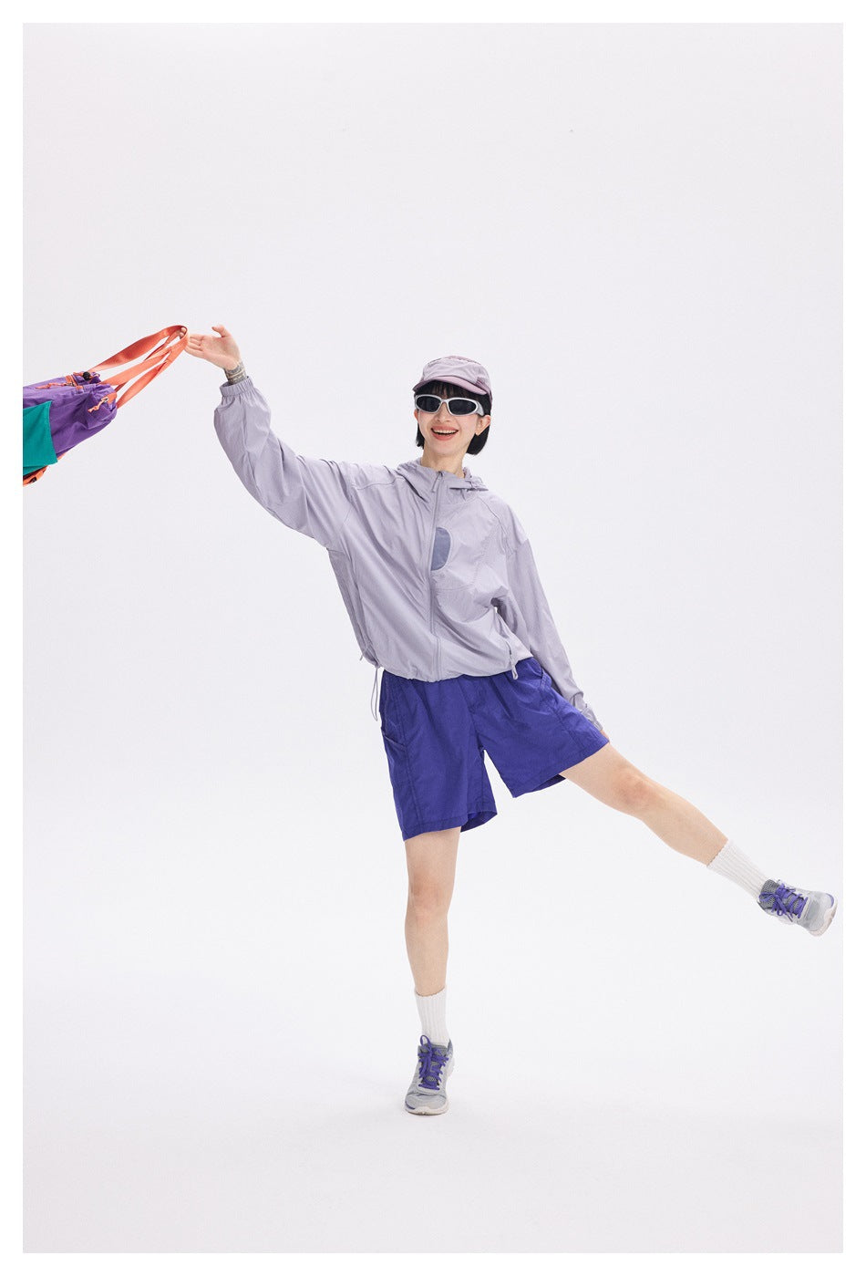 Model in a light lavender hooded jacket and royal blue shorts poses with one leg raised, wearing a cap and sunglasses.