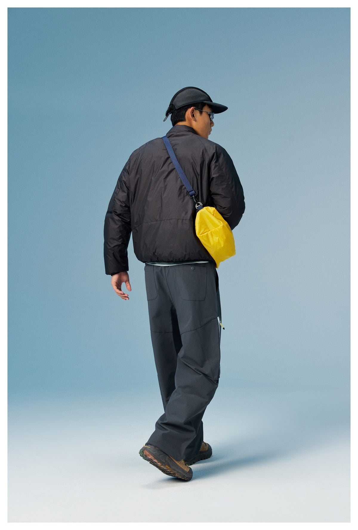 Back view of a model wearing the black jacket with gray pants and a yellow crossbody bag, walking