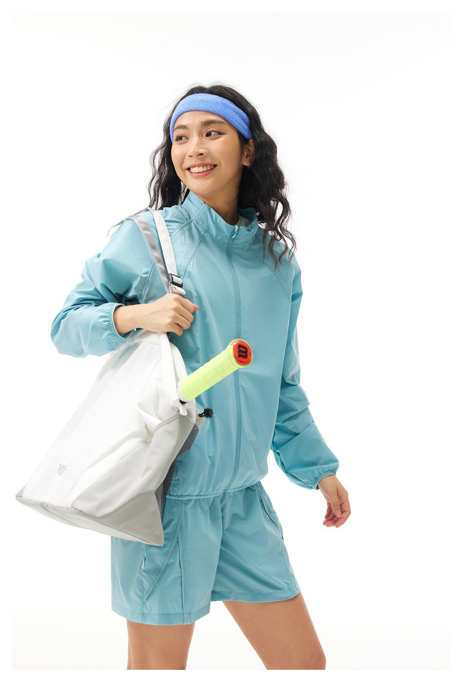 Model in a light blue zip jacket and matching shorts with a blue headband, carrying a white tote bag.