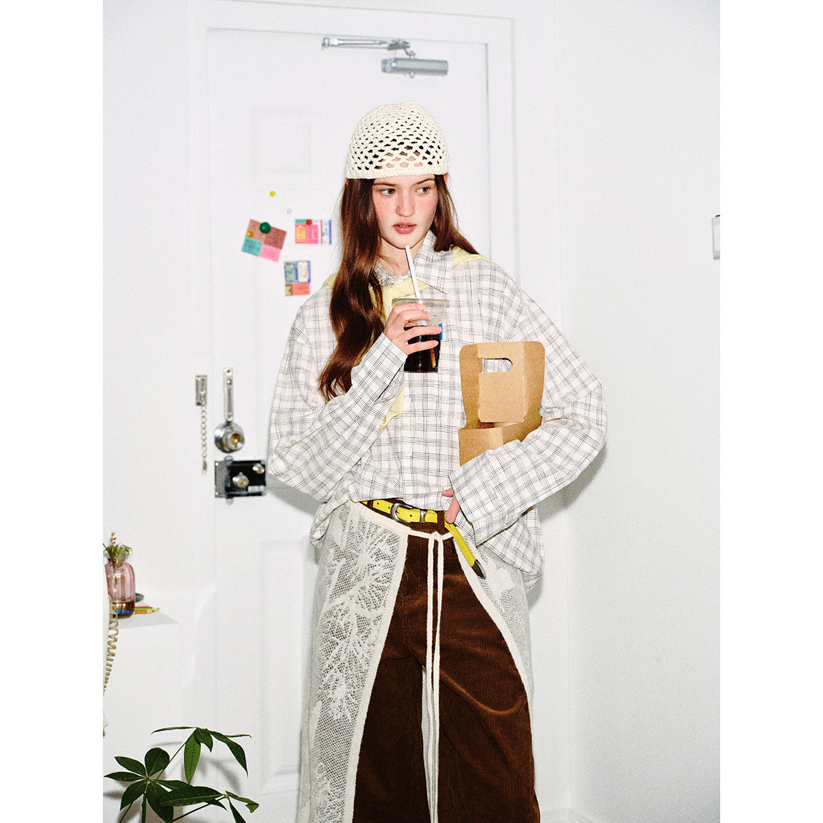 Model in a light plaid shirt and crochet cap, holding a drink; brown pants and a white lace apron are partly visible.