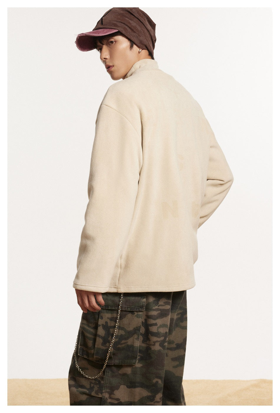 Three-quarter rear view of a model wearing a cream pullover, camouflage cargo pants, and a brown cap.