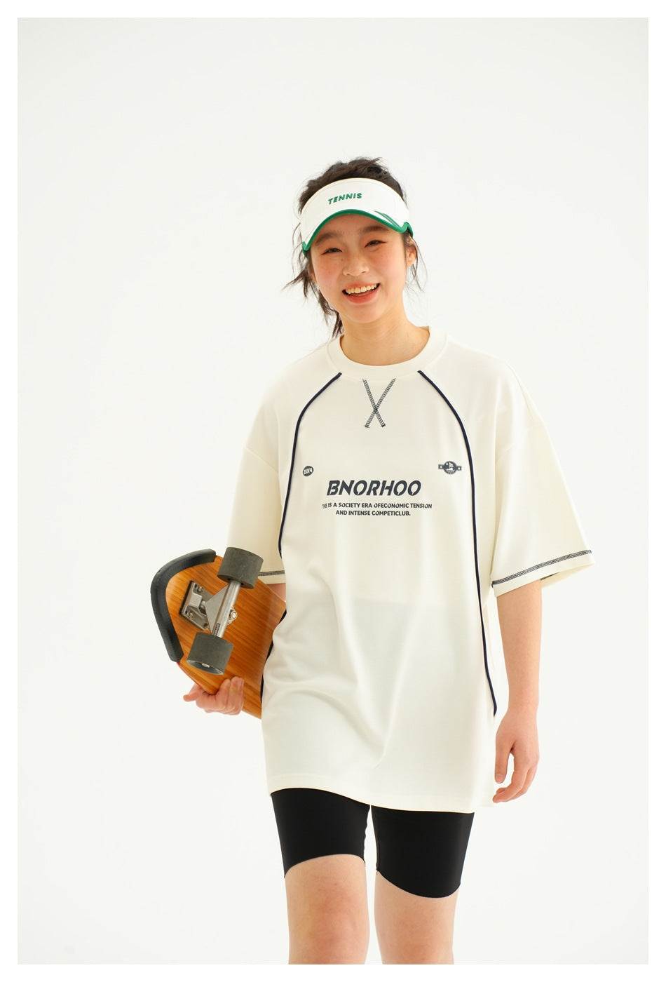 Model wearing an ivory oversized T-shirt with dark piping and chest print, paired with black shorts; skateboard at her side.
