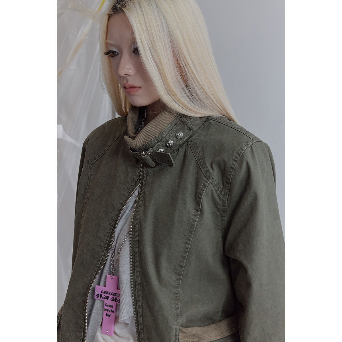 Close-up of a model wearing an olive jacket with a buckled stand collar, silver buttons and a front zipper.
