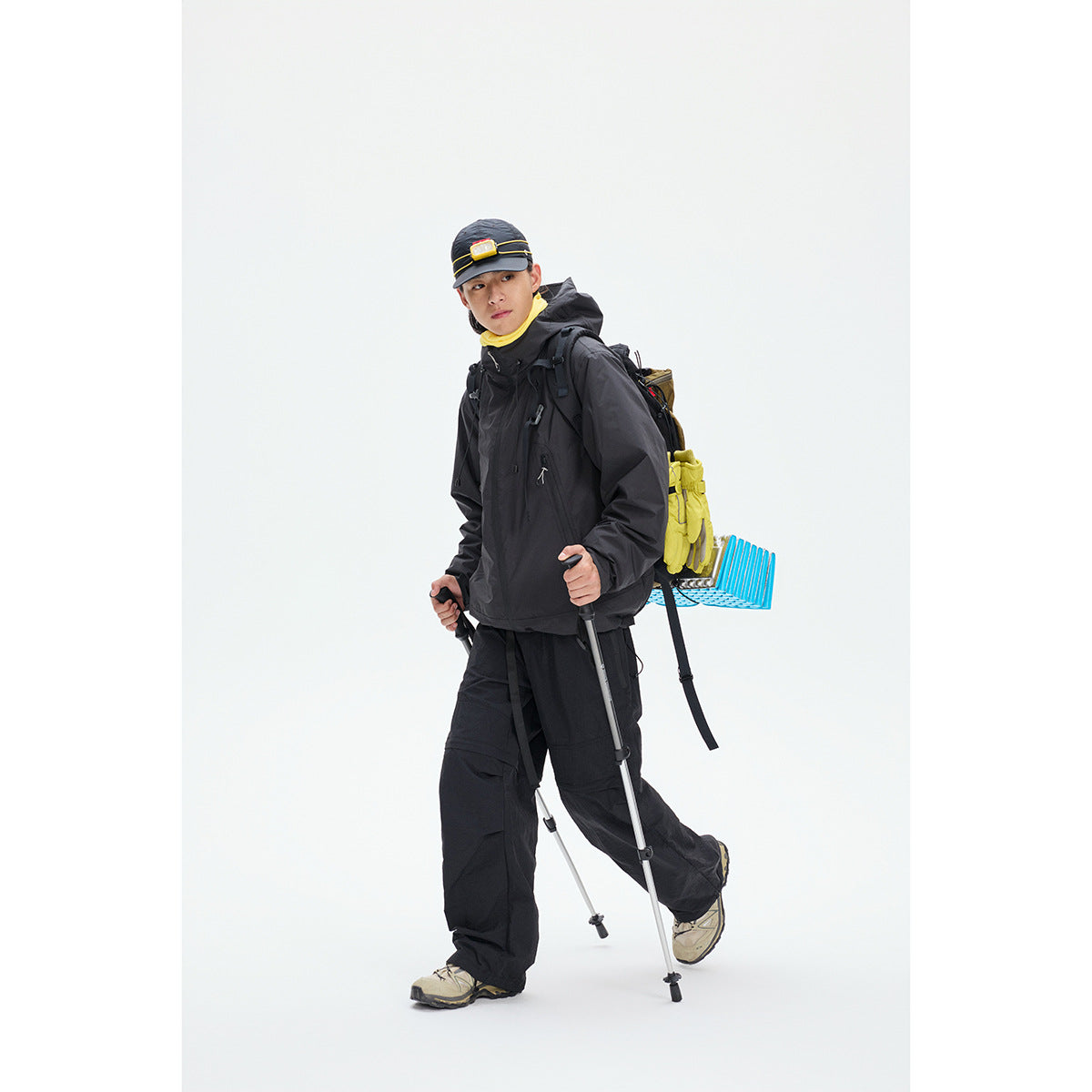 Model wearing a black hooded hiking jacket and pants with a loaded backpack and trekking poles.