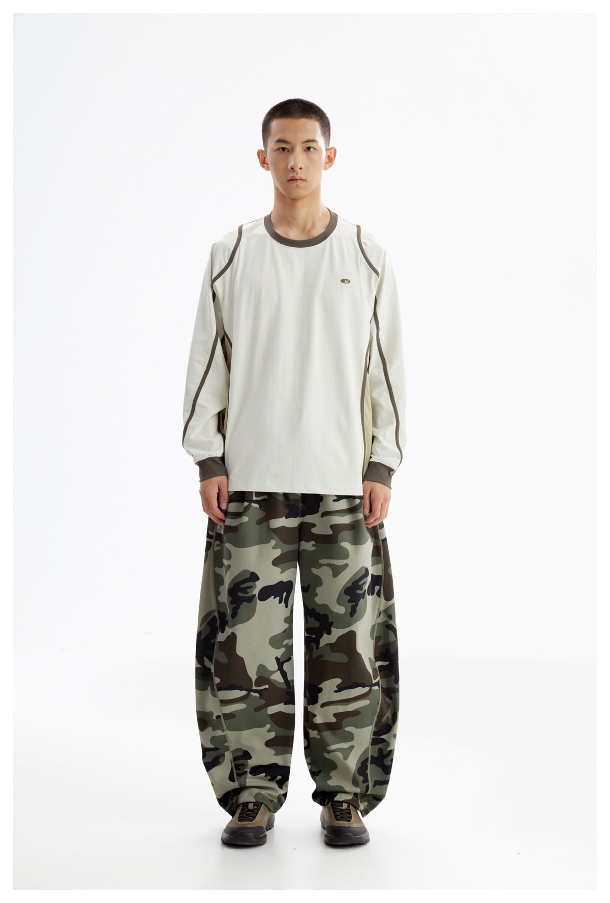 Model wearing an off-white long-sleeve top with dark trim and loose green camouflage trousers, shown from the front.