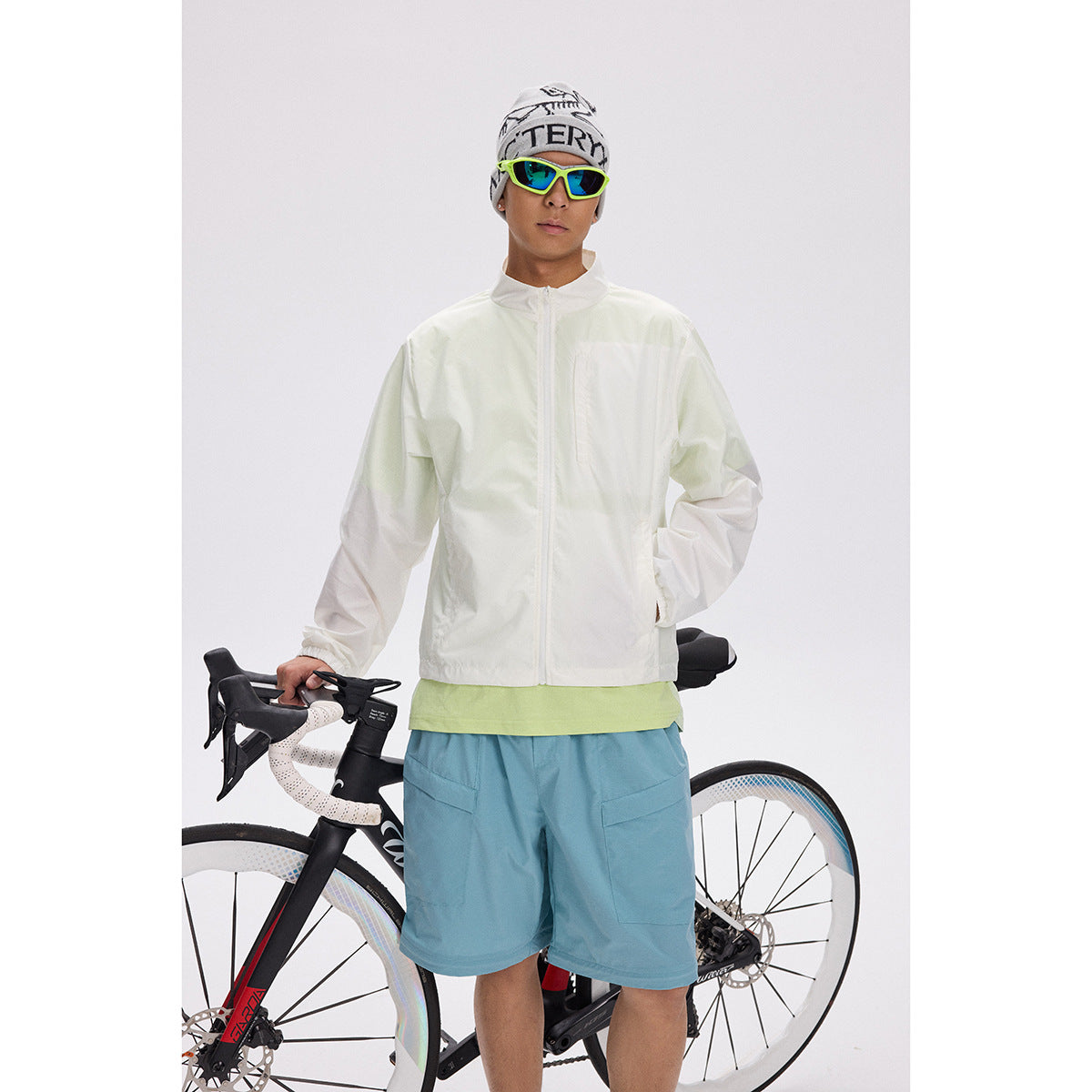 Cyclist wearing a white zip-front jacket and blue shorts, standing with a road bike.