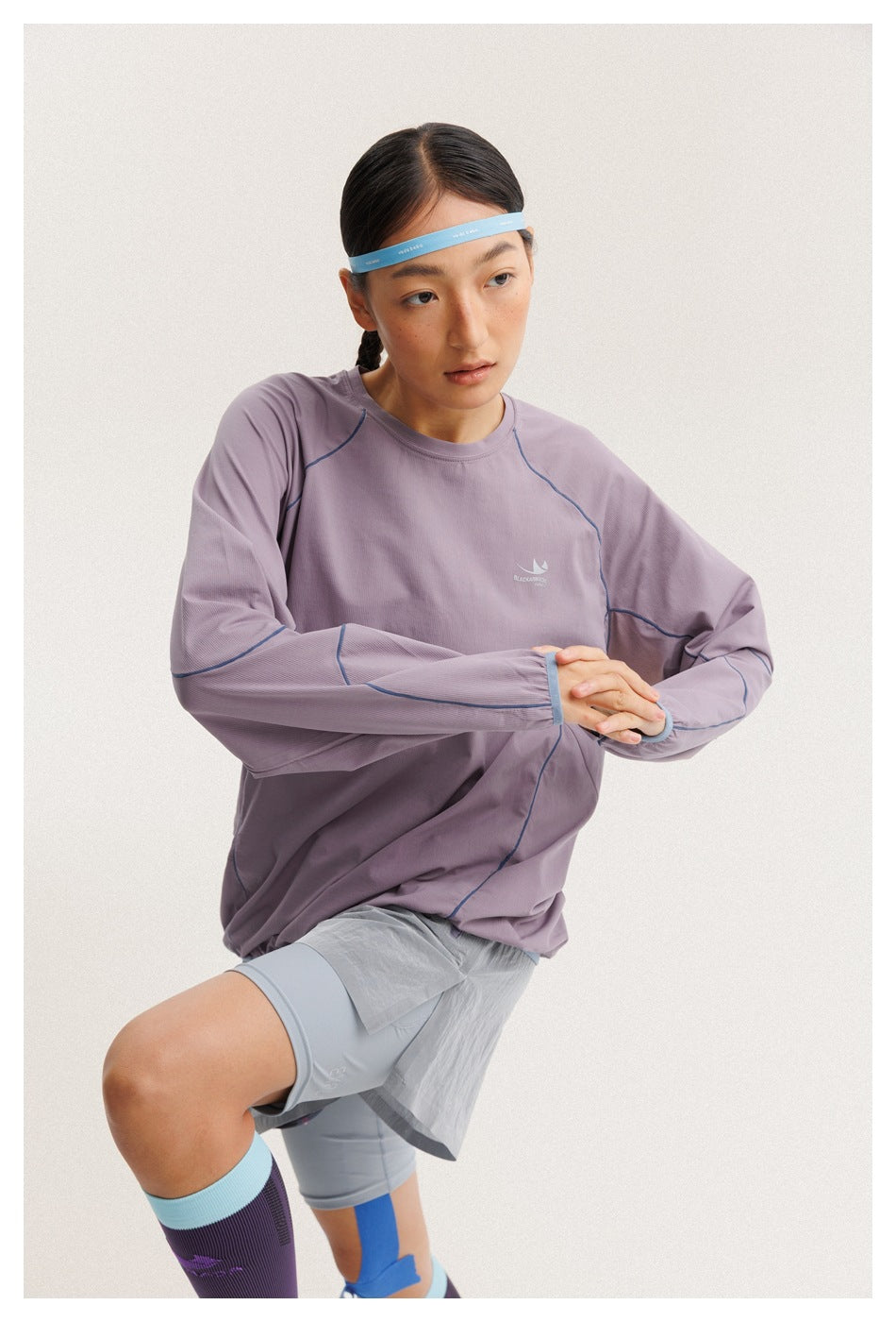 A woman wears a lavender long-sleeve top with blue seam piping; the close view shows its crew neck and chest logo.