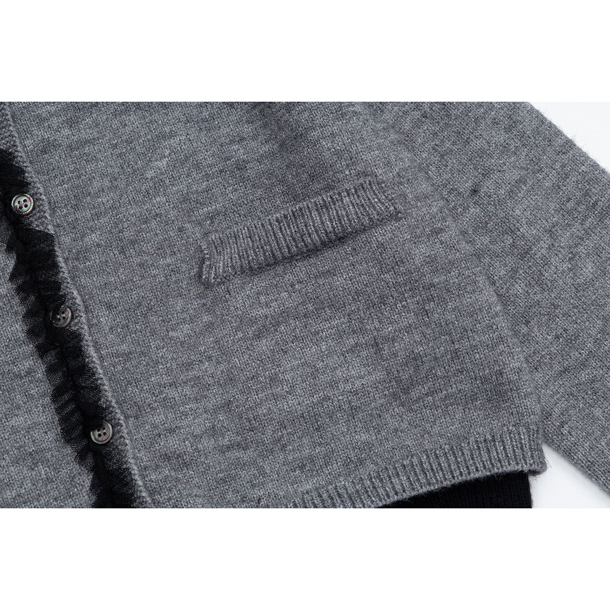 Close-up of a gray knit cardigan showing a ribbed pocket, black ruffled button placket, and contrasting hem.