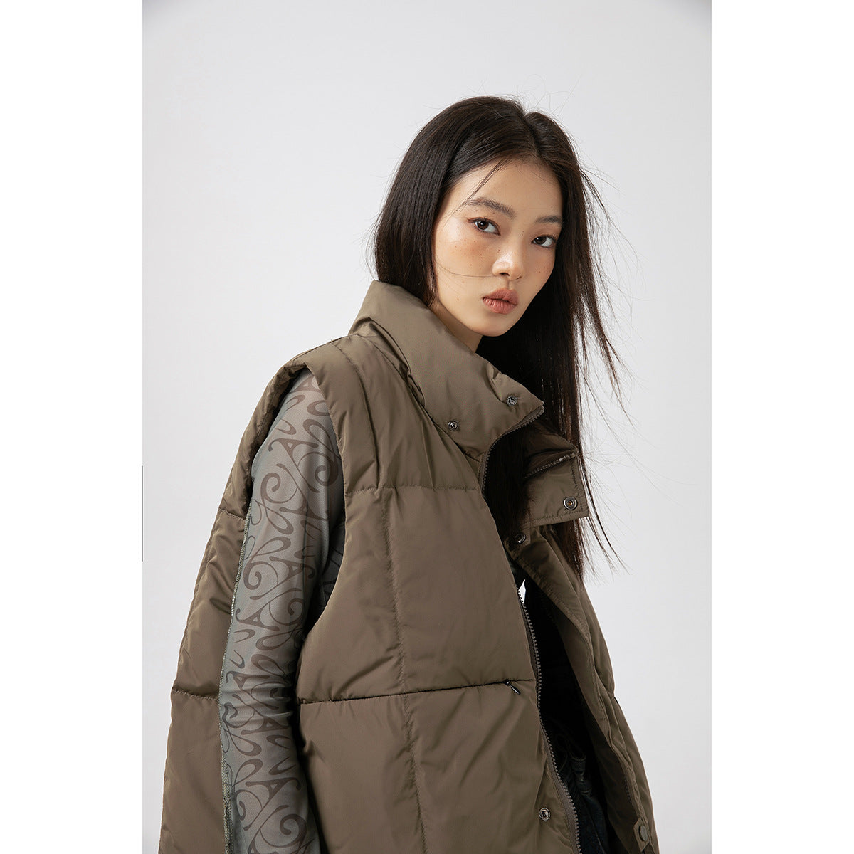 Close-up of a model in a taupe padded vest with a high collar over a gray sheer top with a dark swirling pattern.