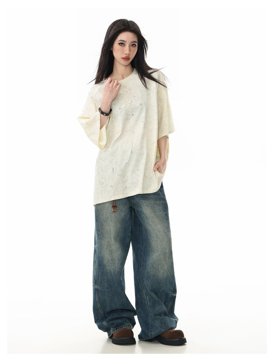 Model wearing an oversized ivory, gray-speckled T-shirt with a small chest emblem and loose dark blue jeans, front view.