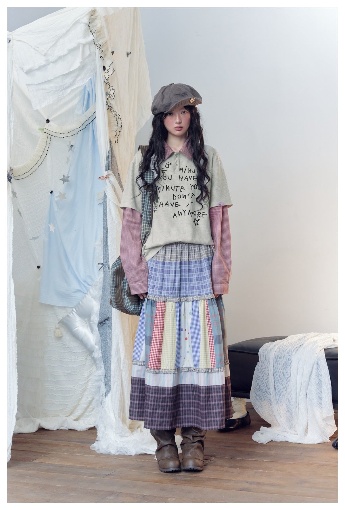 Front view of a model wearing a pale gray graphic tee over a pink checked shirt and a long patchwork plaid skirt.