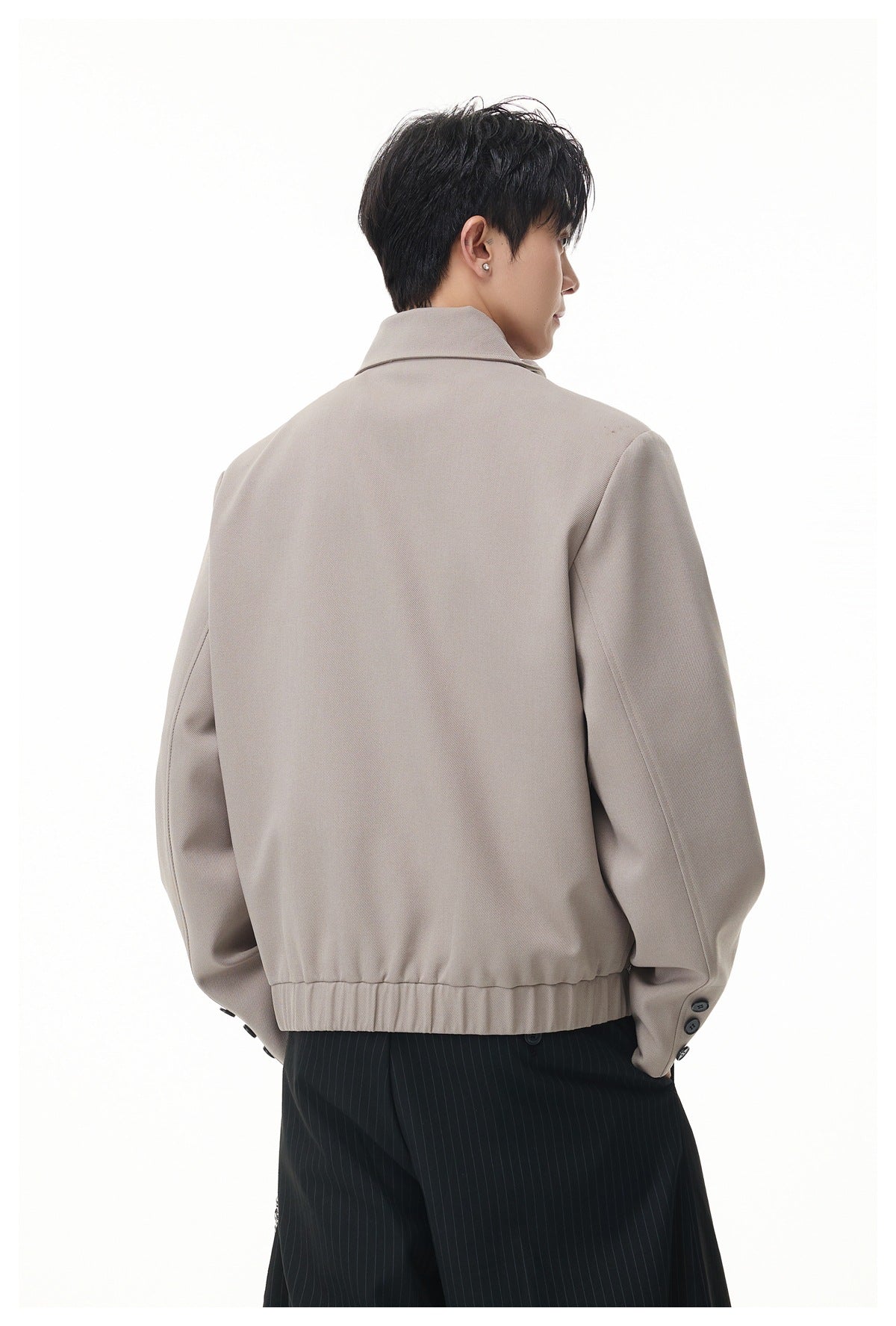 Rear view of a model wearing a taupe jacket with a stand collar, gathered hem and buttoned cuffs.