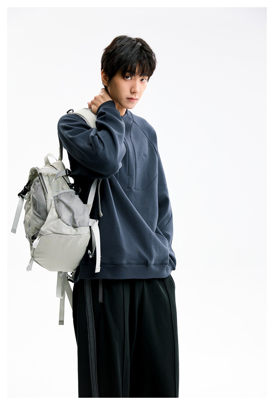 Closer view of a model in a dark navy zip-neck pullover, carrying a light backpack; black pants are partly visible.
