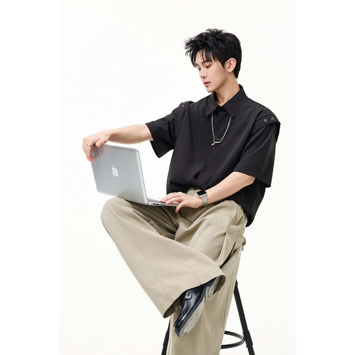 Model seated with a laptop, wearing a black short-sleeve button-up shirt with snap details at the shoulders.