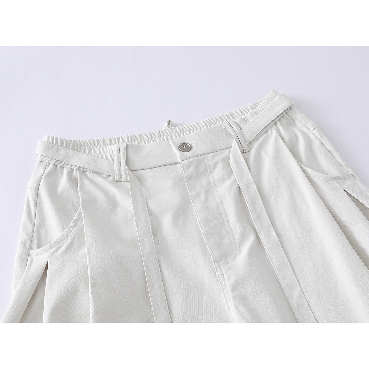Close-up of ivory shorts showing an elasticated waistband, button, belt loops, front pleats, pockets and long belt tabs.