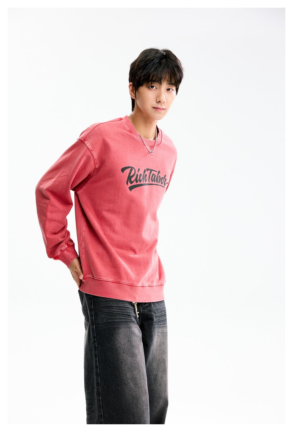 Model wearing a coral-pink crewneck sweatshirt with dark script lettering on the chest.