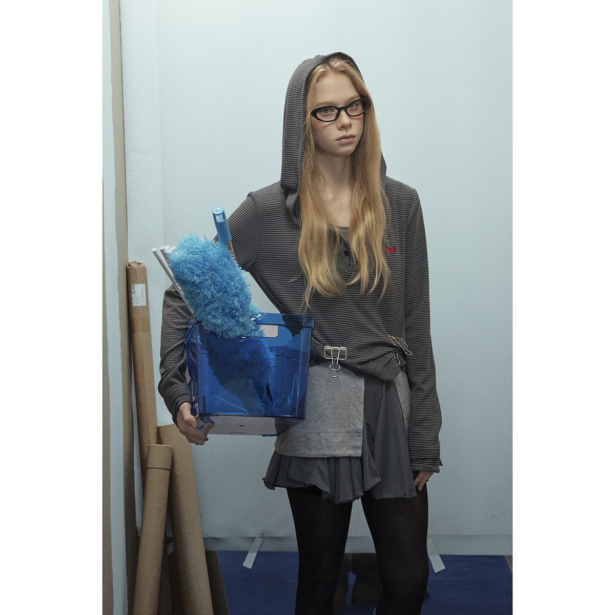 Model wearing a charcoal fine-striped hooded top with a small cherry emblem, layered over a gray top and dark skirt.