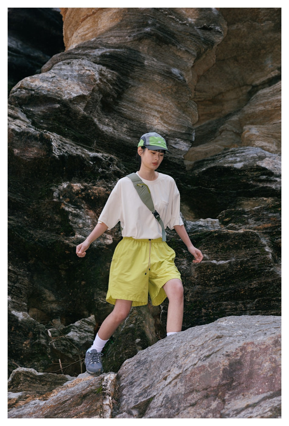 Woman in a white pocket T-shirt, lime-green shorts and cap stepping across rocks outdoors.