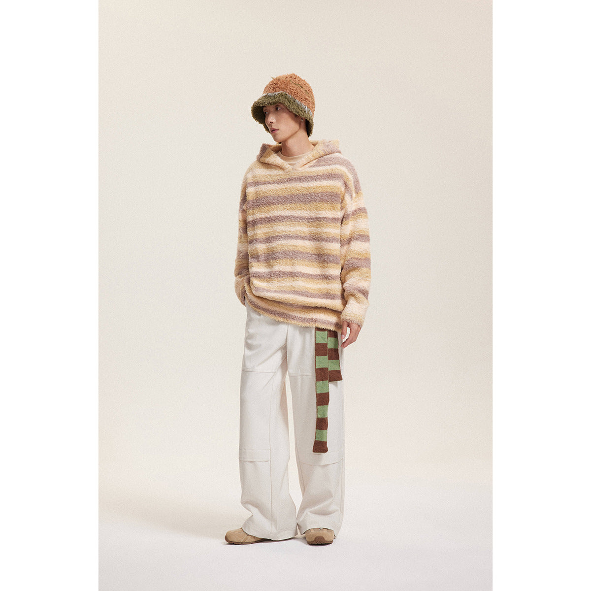 Full-length view of a model in a cream and brown striped hooded sweater, white pants, and a two-tone fuzzy hat.